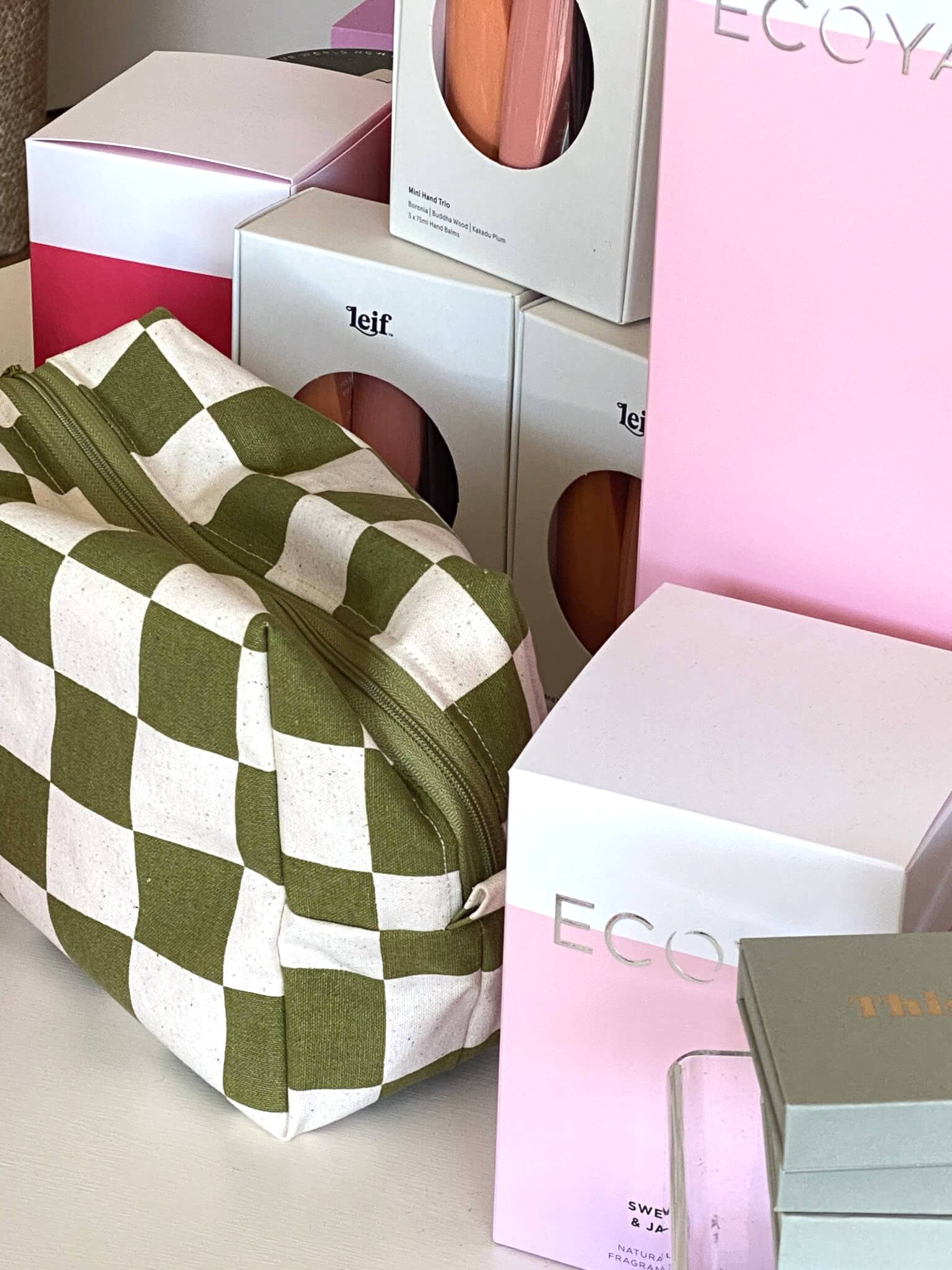 Green checkered pouch displayed alongside Ecoya and Leif product boxes on a white surface, featuring pastel pink and orange tones.