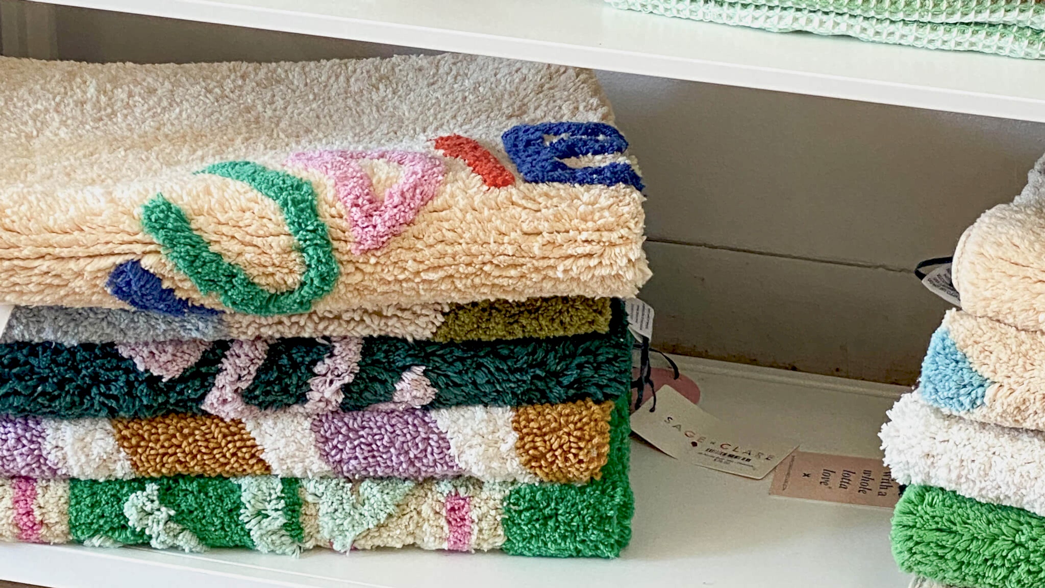 Colorful, patterned Sage + Clare towels neatly stacked on a white shelf, featuring soft textures and vibrant designs.