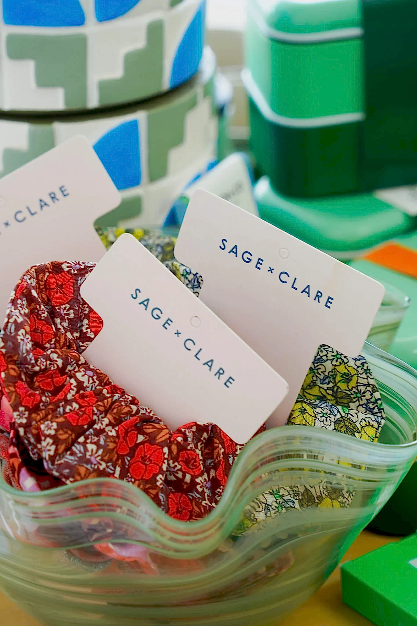 Floral Sage + Clare scrunchies displayed in a green, wavy glass bowl, with colorful boxes and patterns in the background.